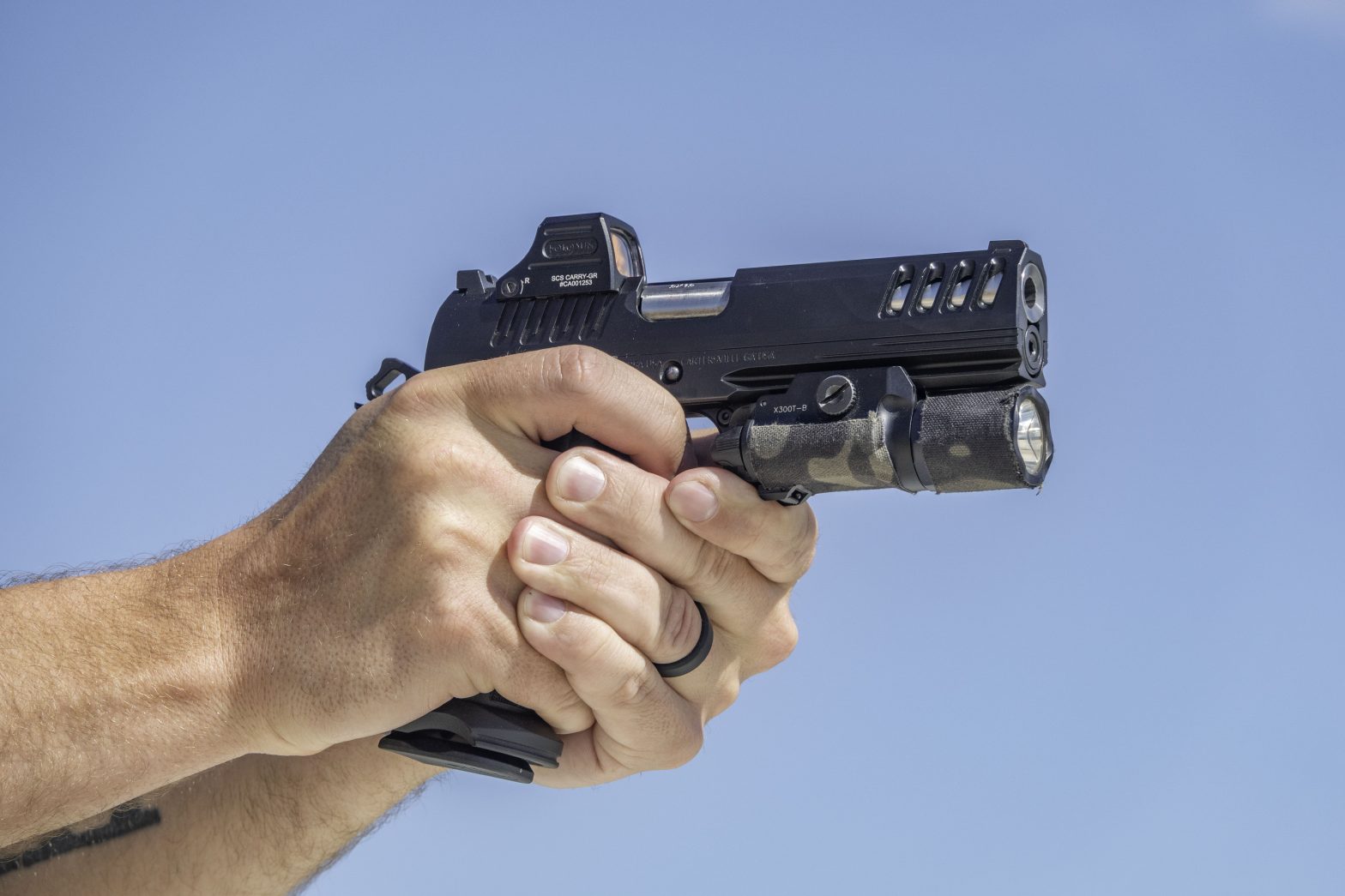 Close-up of two hands gripping the Bersa MX2I black pistol with a weapon light, set against a clear blue sky.